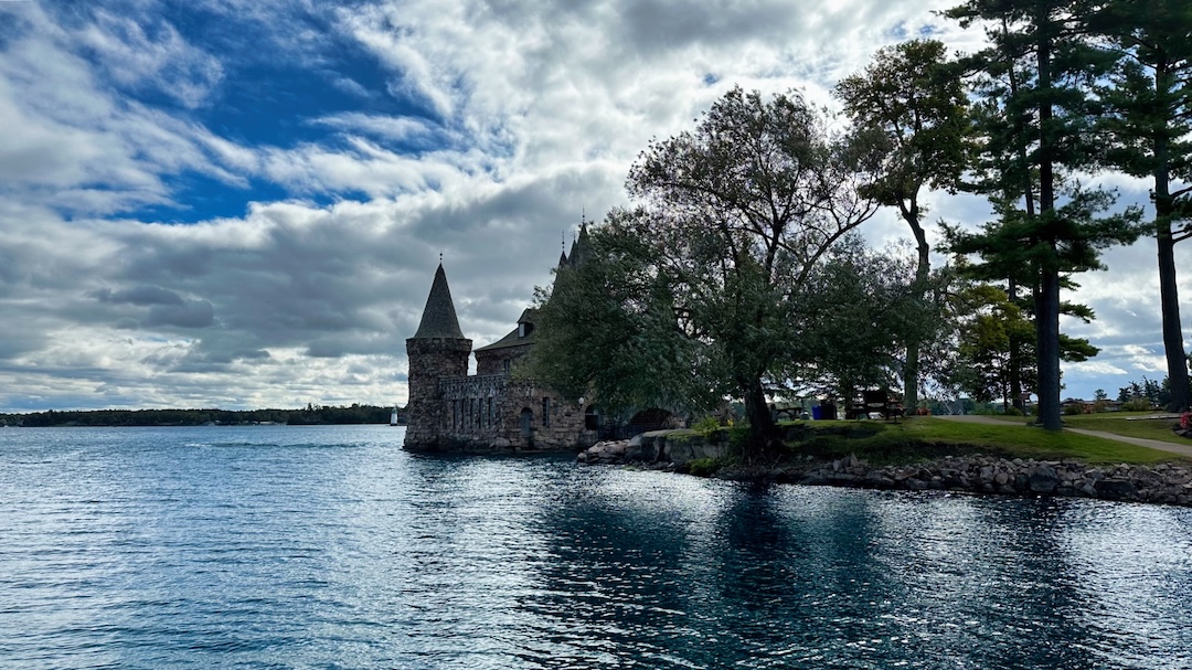 Stone medieval-style power house with a conical tower and slate roofs on the edge of a rocky island shore, with a calm river in the foreground under a dramatic cloudy sky.