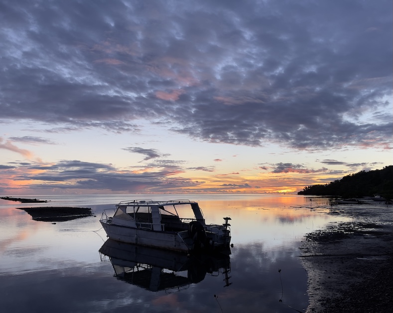 A dive boat moored in calm, shallow water at sunrise, with orange and purple clouds reflected on the glassy bay at Waidroka Bay Resort, Pacific Harbour, Fiji.